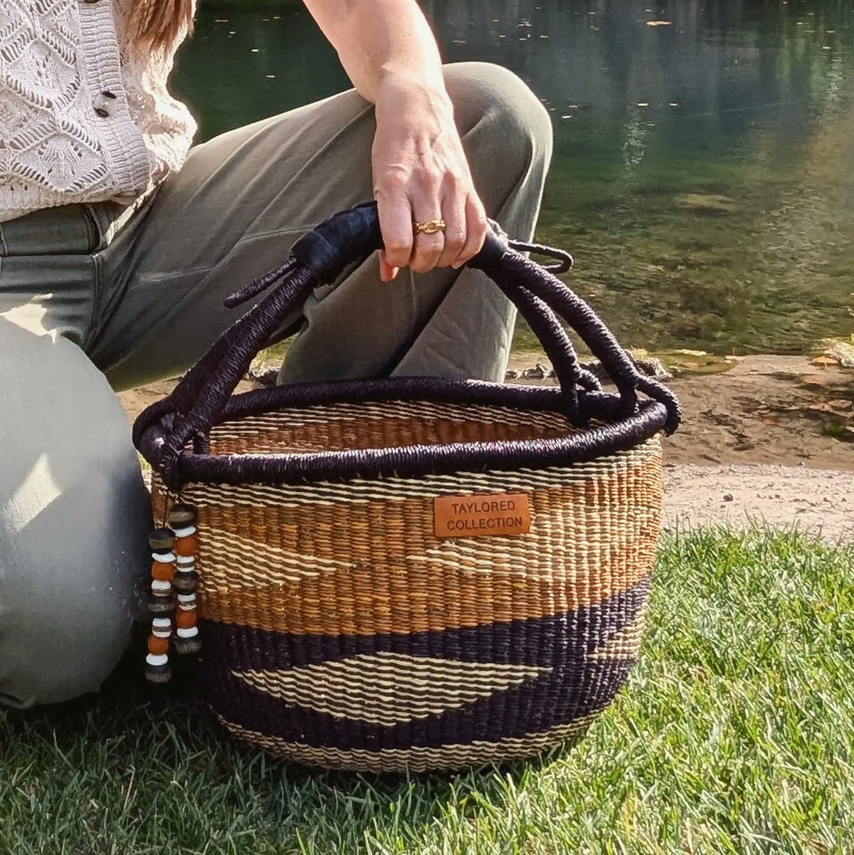 A woven basket bag with dark purple handles and geometric stripes, held by a person sitting outdoors near a body of water. The bag has a tan leather patch reading 'TAYLOR'S COLLECTION'.