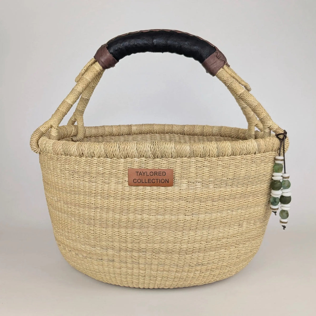 A woven natural straw basket handbag with a black leather handle, brown leather accents, and a small brown leather brand patch reading 'TAYLORED COLLECTION'.