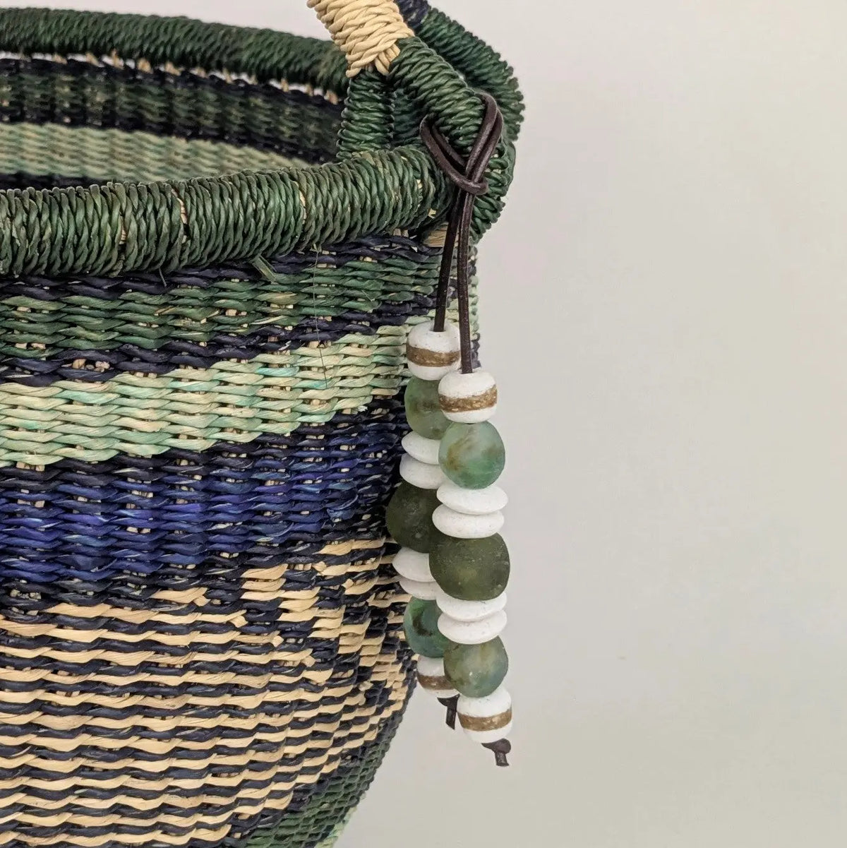 A close-up of a woven basket with green, navy, and beige horizontal stripes, featuring a dark brown leather handle adorned with green and white beaded tassels.