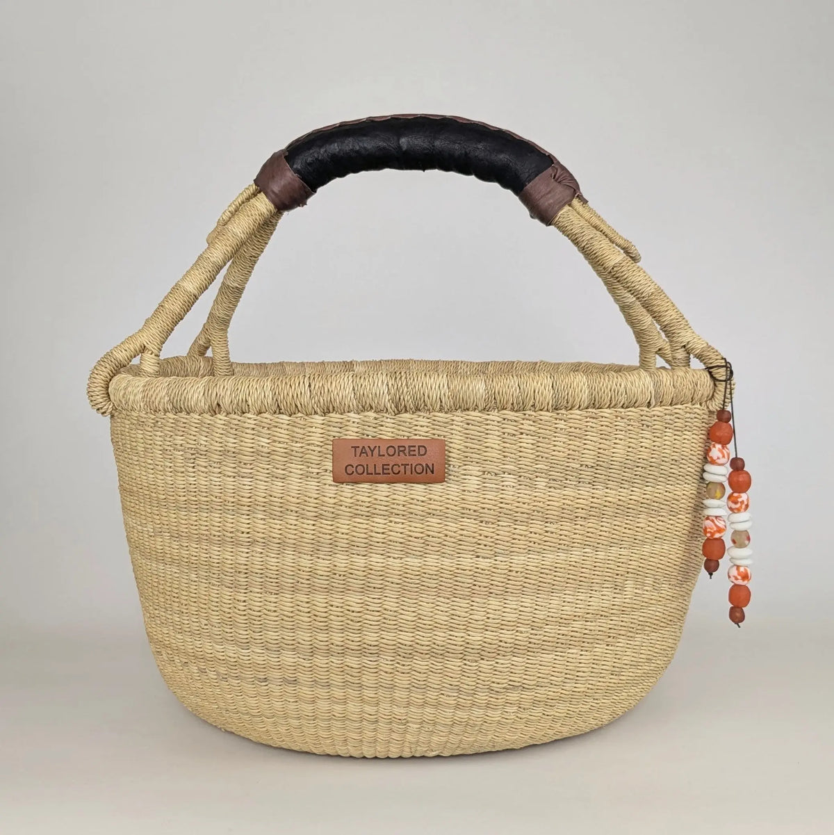 A woven straw basket bag with a curved top handle, featuring a black leather-wrapped handle, a small brown leather patch reading 'TAYLORED COLLECTION', and a dangling beaded charm.