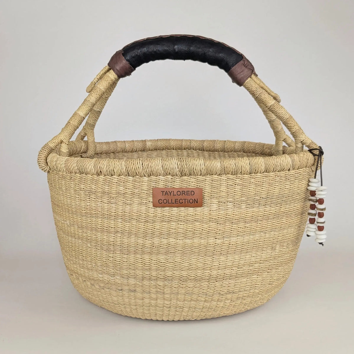 A natural woven straw basket handbag with a dark brown leather handle and a small brown leather brand patch reading 'TAYLORED COLLECTION'.