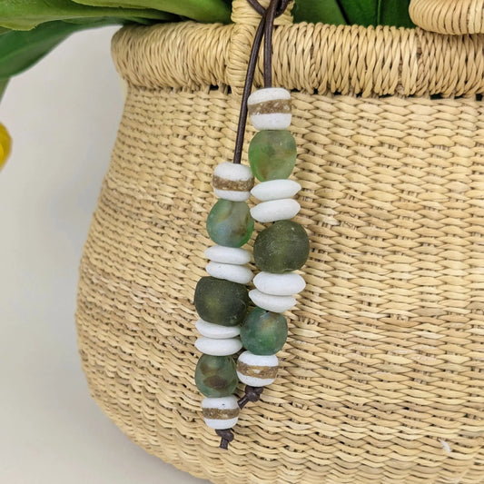 A beaded hanging charm or ornament with round green and white beads, suspended from a woven natural fiber basket with green leaves visible above.