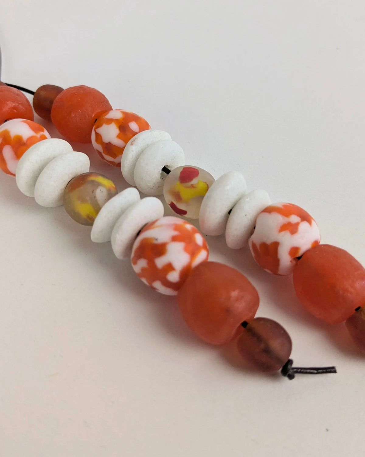 A close-up of a beaded strand featuring orange and white patterned beads, translucent orange beads, and white textured discs on a light beige background.