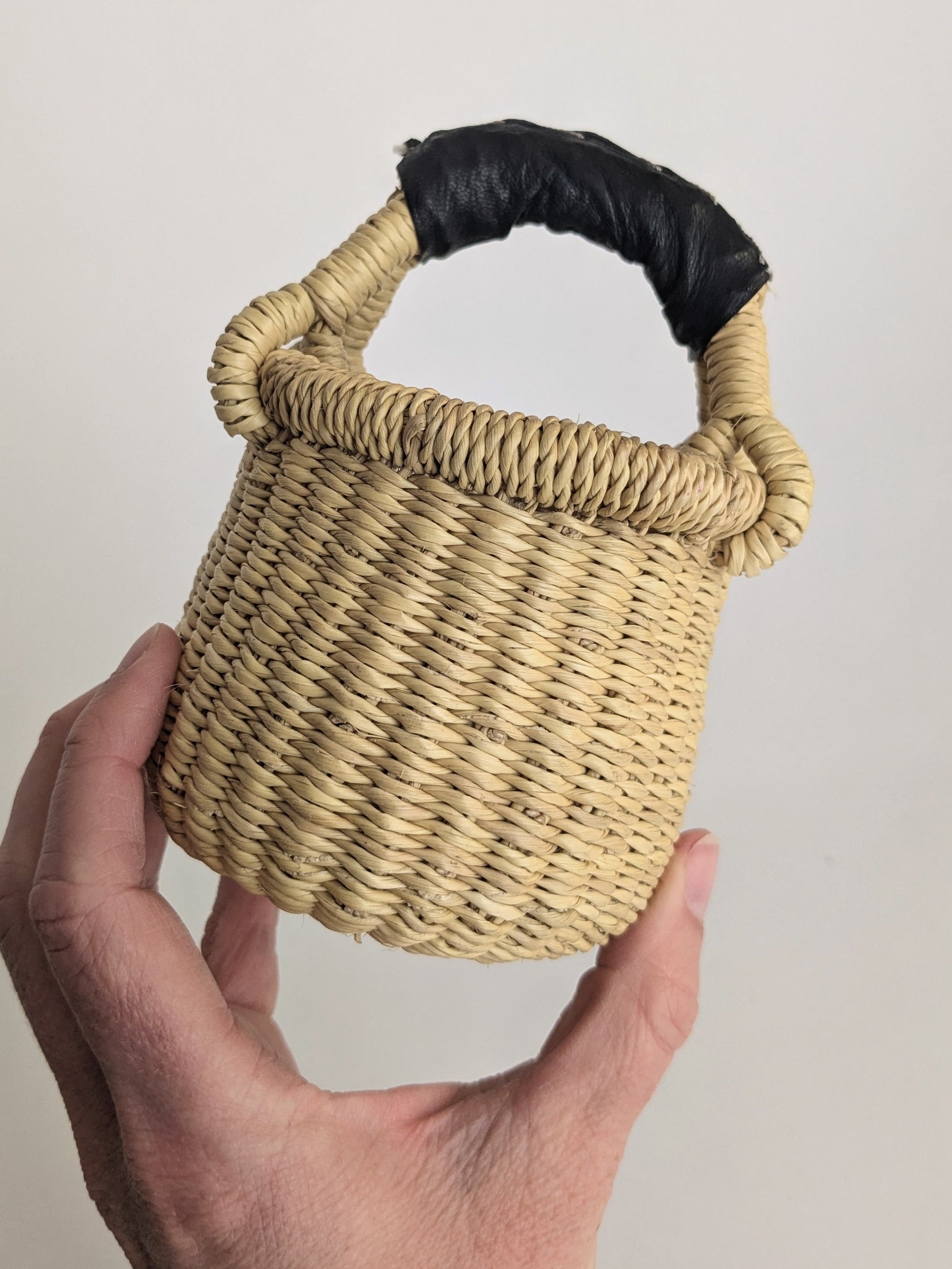 Hand holding a small woven basket with a black handle against a plain background