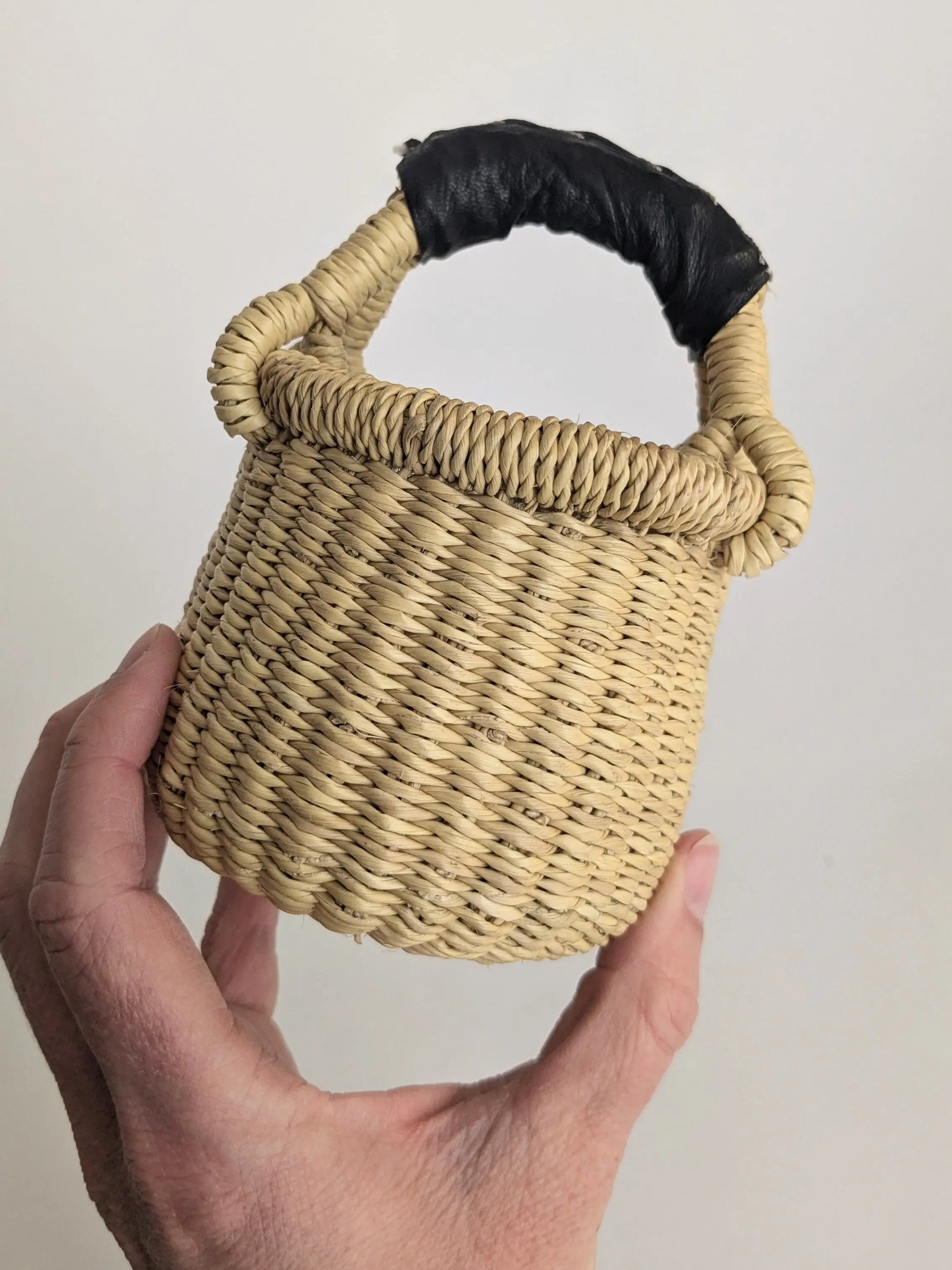 Hand holding a small woven basket with a black handle against a plain background