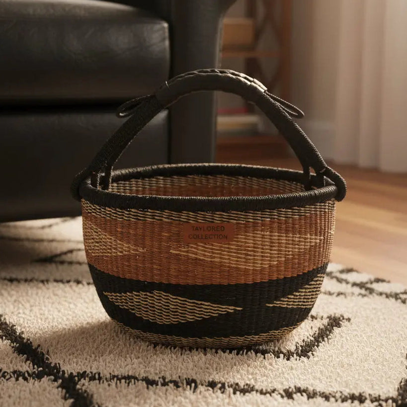 A woven basket with a black leather handle and trim, featuring natural tan and black geometric stripes, sitting on a patterned rug next to a black leather sofa.